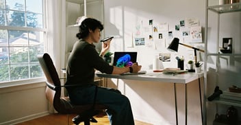 Person sitting at a desk working on a laptop, holding a phone, with papers, photos, and notes pinned to the wall in a well-lit room with a large window.